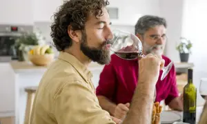 Mann genießt ein Glas Rotwein am Abend, während ein Freund im Hintergrund sitzt.