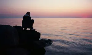 Ein Mann hockt auf Felsen am Meer, blickt in die Ferne. Der Himmel ist in sanften Rosa- und Orangetönen gefärbt, während das Wasser ruhig glitzert. Stille Gedanken in der Abenddämmerung.