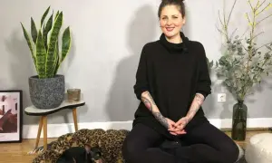 Frau in schwarzem Pullover sitzt im Lotussitz, bereit für eine herzmeditation. Ein kleiner Hund ruht auf einem Leopardenmuster-Bett.