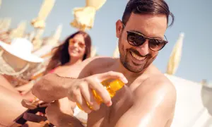 Mann sprüht sich Sonnencreme auf den Arm, um Sonnenbrand vorzubeugen. Im Hintergrund entspannt eine Frau im Liegestuhl.