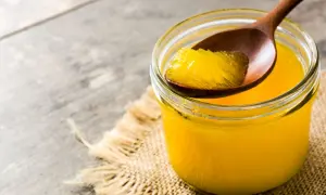 Ein Glas mit goldgelbem Ghee, das auf einem Holzlöffel entnommen wird. Ghee ist geklärte Butter, ein vielseitiges Fett.