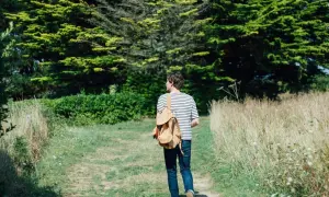 Mann mit Rucksack auf einem Feldweg, umgeben von grünen Bäumen und Gräsern. Ein Moment des meditativen Gehens in der Natur.