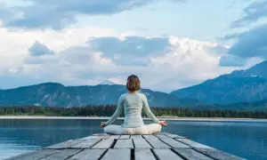 Frau in Lotusposition auf einem Steg, genießt eine einfache Meditation mit Blick auf einen Bergsee und die Alpen.