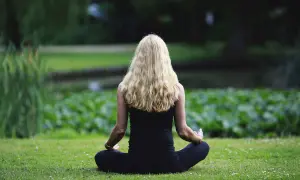Frau mit langen blonden Haaren meditiert im Lotussitz im Grünen. Sie strahlt Geduld und Gelassenheit aus.