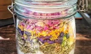 Ein Glas mit selbstgemachtem Badezusatz: unten Salz, darauf Rosmarin, dann lila Blüten und oben rosa Blütenblätter.