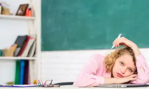 Junge Lehrerin mit Locken, die mit einem Stift auf dem Schoß liegt, wirkt gestresst. Hinter ihr eine Tafel und ein Regal voller Bücher.