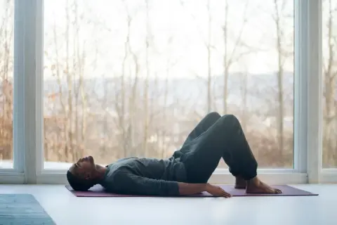 Mann liegt auf Yogamatte, Knie angewinkelt, entspannt. Fokus auf tiefe meditation atmung vor großem Fenster mit Blick auf kahle Bäume.