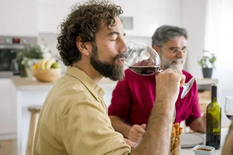 Mann genießt ein Glas Rotwein am Abend, während ein Freund im Hintergrund sitzt.