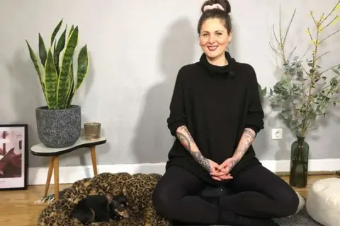 Frau in schwarzem Pullover sitzt im Lotussitz, bereit für eine herzmeditation. Ein kleiner Hund ruht auf einem Leopardenmuster-Bett.
