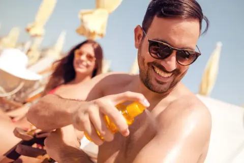Mann sprüht sich Sonnencreme auf den Arm, um Sonnenbrand vorzubeugen. Im Hintergrund entspannt eine Frau im Liegestuhl.