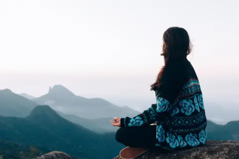 Frau meditiert auf einem Felsen mit Blick auf nebelverhangene Berge. Sie übt schweigen statt reden, um innere Ruhe zu finden.