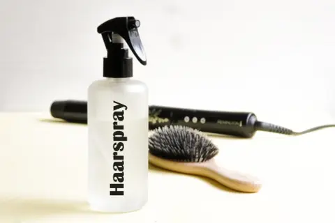 Flasche mit "Haarspray" Schriftzug, daneben Haarbürste und Glätteisen. Perfekt für haarspray selber machen.