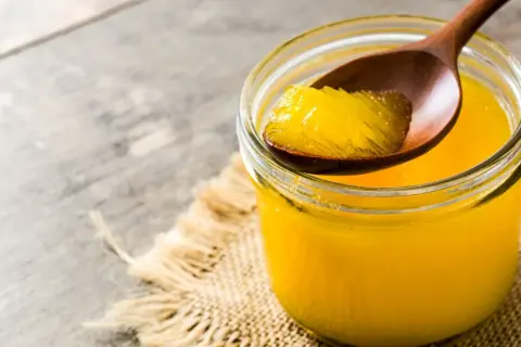 Ein Glas mit goldgelbem Ghee, das auf einem Holzlöffel entnommen wird. Ghee ist geklärte Butter, ein vielseitiges Fett.