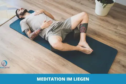 Mann mit Tattoos entspannt sich im Liegen auf einer Yogamatte, umgeben von Holzboden und einer Pflanze. Perfekt für Meditation im Liegen.