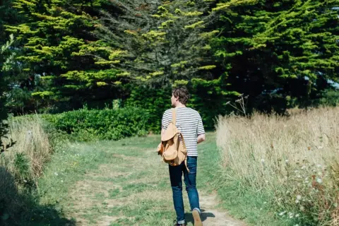 Mann mit Rucksack auf einem Feldweg, umgeben von grünen Bäumen und Gräsern. Ein Moment des meditativen Gehens in der Natur.