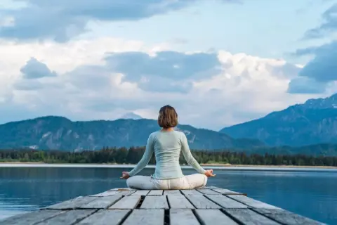 Frau in Lotusposition auf einem Steg, genießt eine einfache Meditation mit Blick auf einen Bergsee und die Alpen.