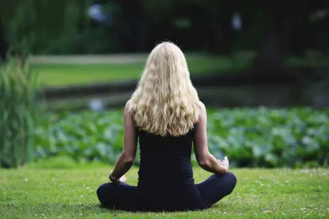 Frau mit langen blonden Haaren meditiert im Lotussitz im Grünen. Sie strahlt Geduld und Gelassenheit aus.