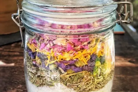 Ein Glas mit selbstgemachtem Badezusatz: unten Salz, darauf Rosmarin, dann lila Blüten und oben rosa Blütenblätter.
