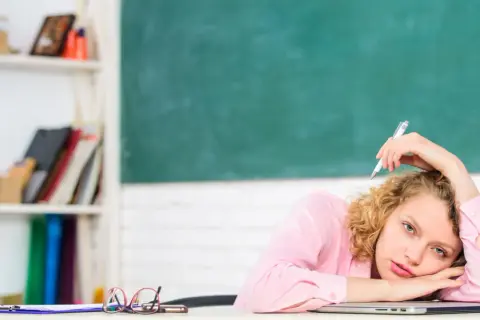 Junge Lehrerin mit Locken, die mit einem Stift auf dem Schoß liegt, wirkt gestresst. Hinter ihr eine Tafel und ein Regal voller Bücher.