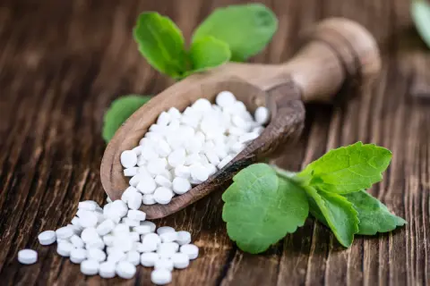 Süße Tabletten aus Stevia auf einem Holzlöffel, umgeben von grünen Blättern. Mögliche stevia nachteile sind hier nicht ersichtlich.