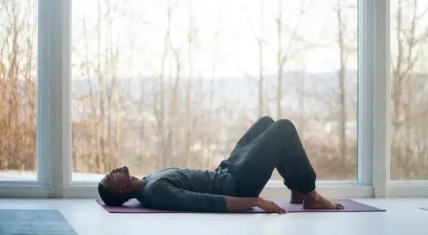 Mann liegt auf Yogamatte, Knie angewinkelt, entspannt. Fokus auf tiefe meditation atmung vor großem Fenster mit Blick auf kahle Bäume.