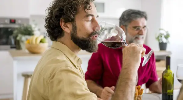 Mann genießt ein Glas Rotwein am Abend, während ein Freund im Hintergrund sitzt.