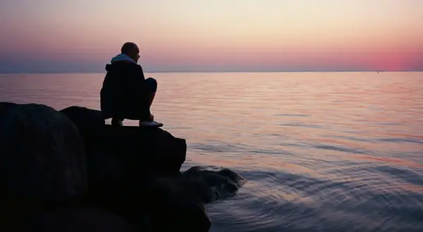 Ein Mann hockt auf Felsen am Meer, blickt in die Ferne. Der Himmel ist in sanften Rosa- und Orangetönen gefärbt, während das Wasser ruhig glitzert. Stille Gedanken in der Abenddämmerung.