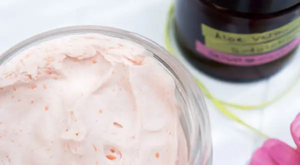 Cremige, rosafarbene Bodylotion mit Sheabutter in einem Glas. Daneben eine Flasche mit "Aloe Vera Bodylotion".