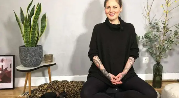 Frau in schwarzem Pullover sitzt im Lotussitz, bereit für eine herzmeditation. Ein kleiner Hund ruht auf einem Leopardenmuster-Bett.
