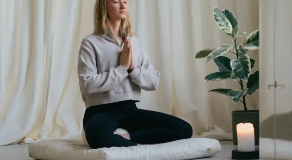 Frau in Meditation, die auf einem Kissen sitzt und mit geschlossenen Augen und gefalteten Händen zur Ruhe kommt. Sie übt, Sorgen loszulassen.
