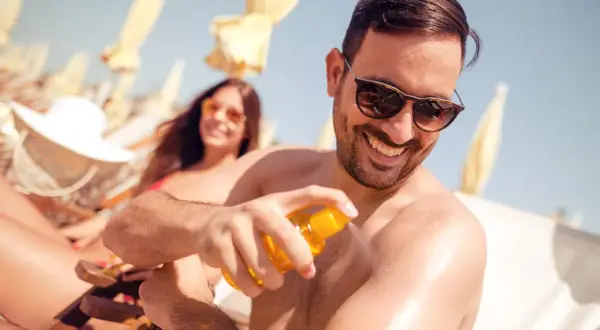 Mann sprüht sich Sonnencreme auf den Arm, um Sonnenbrand vorzubeugen. Im Hintergrund entspannt eine Frau im Liegestuhl.