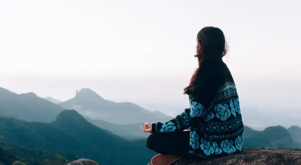 Frau meditiert auf einem Felsen mit Blick auf nebelverhangene Berge. Sie übt schweigen statt reden, um innere Ruhe zu finden.