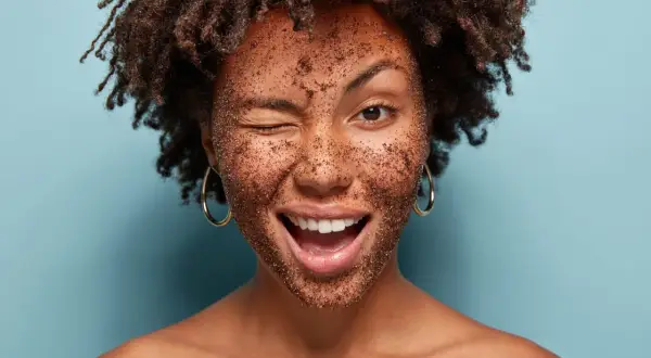 Frau mit braunem Peeling im Gesicht, die zwinkert und lacht. Ein tolles Beispiel, wie man ein Heilerde Peeling selber machen kann.