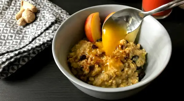 Haferflocken mit Apfelstücken und Rosinen, übergossen mit Honig. Ein leckeres Frühstück, inspiriert von TCM Rezepten. Ingwer liegt daneben.