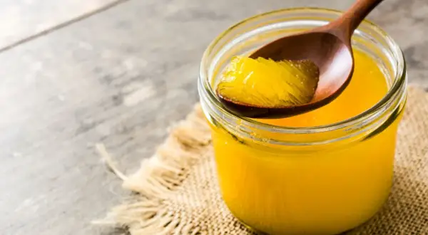 Ein Glas mit goldgelbem Ghee, das auf einem Holzlöffel entnommen wird. Ghee ist geklärte Butter, ein vielseitiges Fett.