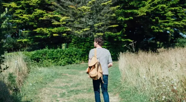 Mann mit Rucksack auf einem Feldweg, umgeben von grünen Bäumen und Gräsern. Ein Moment des meditativen Gehens in der Natur.