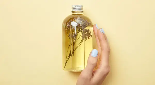 Eine Hand mit hellblauen Nägeln berührt eine Flasche mit gelbem Öl und getrockneten Blumen. Perfekt für duschgel selber machen ohne seife.