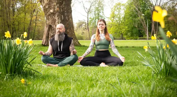 Mann und Frau in bequemer Kleidung machen Meditation im Sitzen auf einer Wiese, umgeben von Narzissen und Bäumen.