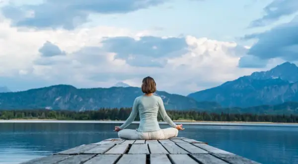 Frau in Lotusposition auf einem Steg, genießt eine einfache Meditation mit Blick auf einen Bergsee und die Alpen.