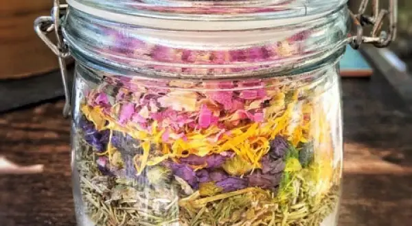 Ein Glas mit selbstgemachtem Badezusatz: unten Salz, darauf Rosmarin, dann lila Blüten und oben rosa Blütenblätter.