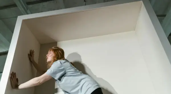 Eine Frau mit roten Haaren stemmt sich gegen eine Wand, als wolle sie die Opferrolle verlassen und aus einer Begrenzung ausbrechen.