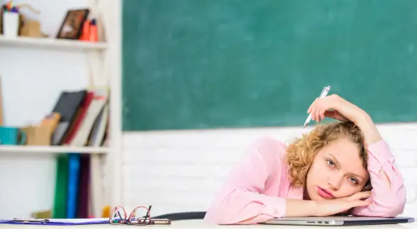 Junge Lehrerin mit Locken, die mit einem Stift auf dem Schoß liegt, wirkt gestresst. Hinter ihr eine Tafel und ein Regal voller Bücher.