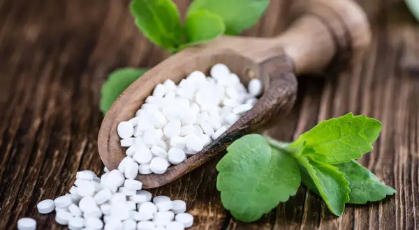 Süße Tabletten aus Stevia auf einem Holzlöffel, umgeben von grünen Blättern. Mögliche stevia nachteile sind hier nicht ersichtlich.