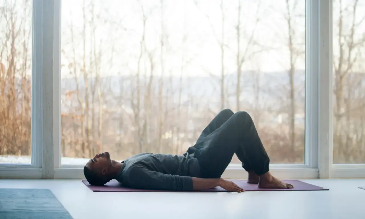 Mann liegt auf Yogamatte, Knie angewinkelt, entspannt. Fokus auf tiefe meditation atmung vor großem Fenster mit Blick auf kahle Bäume.