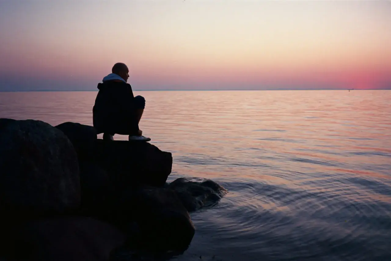 Ein Mann hockt auf Felsen am Meer, blickt in die Ferne. Der Himmel ist in sanften Rosa- und Orangetönen gefärbt, während das Wasser ruhig glitzert. Stille Gedanken in der Abenddämmerung.