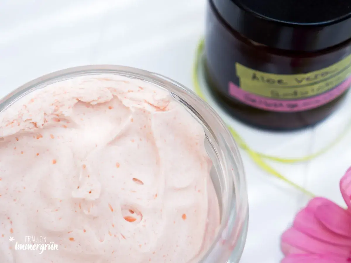 Cremige, rosafarbene Bodylotion mit Sheabutter in einem Glas. Daneben eine Flasche mit "Aloe Vera Bodylotion".