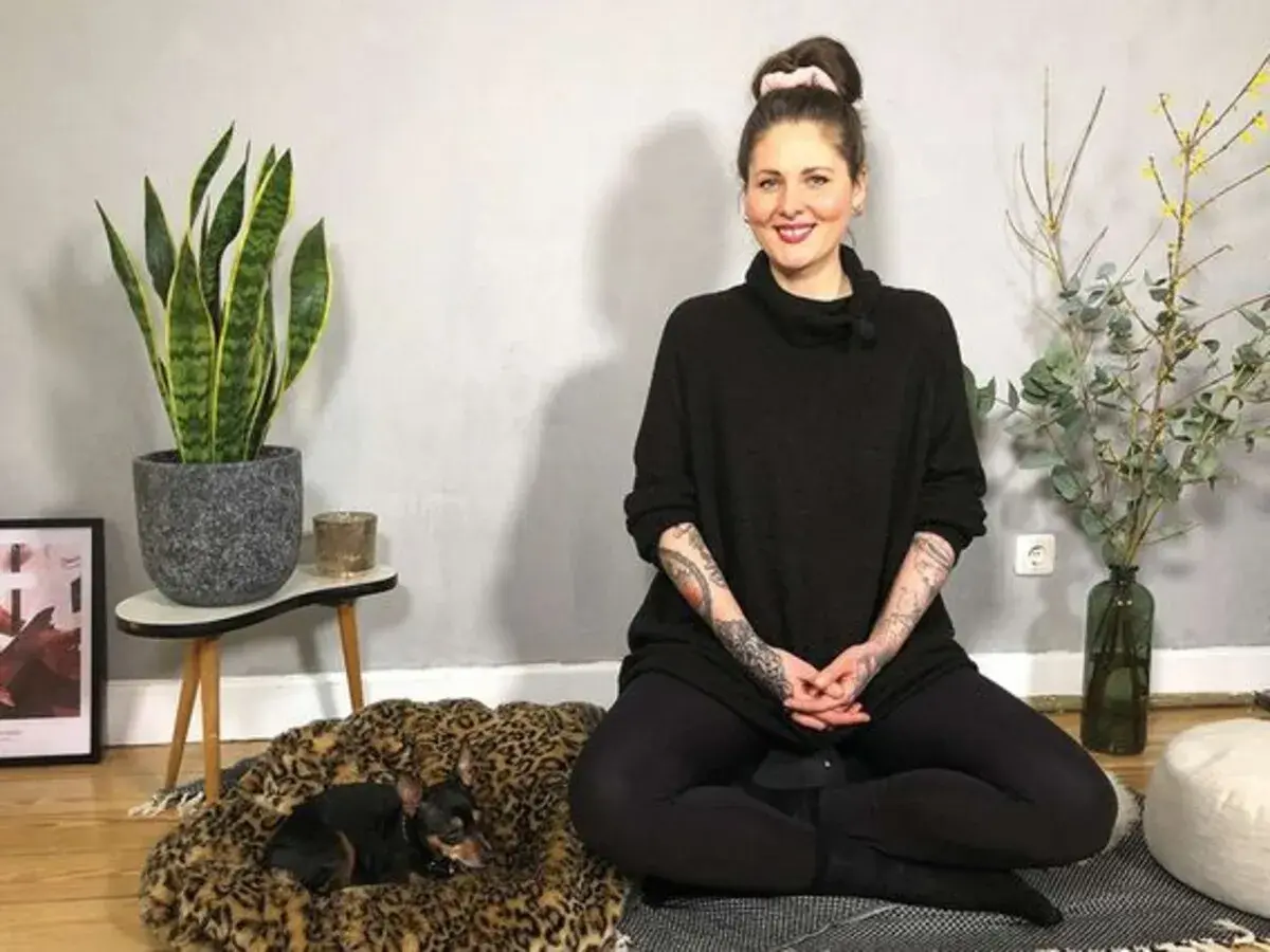 Frau in schwarzem Pullover sitzt im Lotussitz, bereit für eine herzmeditation. Ein kleiner Hund ruht auf einem Leopardenmuster-Bett.