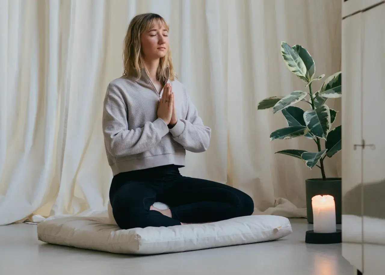 Frau in Meditation, die auf einem Kissen sitzt und mit geschlossenen Augen und gefalteten Händen zur Ruhe kommt. Sie übt, Sorgen loszulassen.