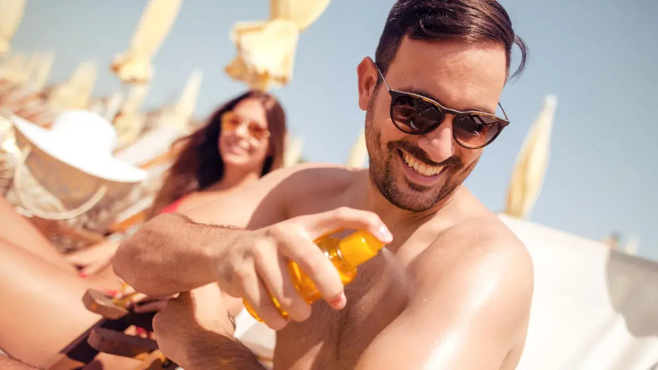 Mann sprüht sich Sonnencreme auf den Arm, um Sonnenbrand vorzubeugen. Im Hintergrund entspannt eine Frau im Liegestuhl.
