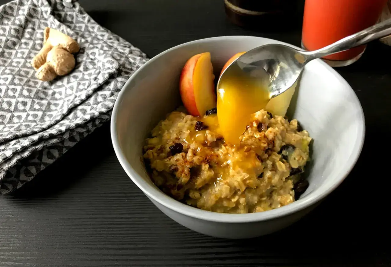 Haferflocken mit Apfelstücken und Rosinen, übergossen mit Honig. Ein leckeres Frühstück, inspiriert von TCM Rezepten. Ingwer liegt daneben.